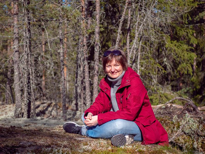 Keski-ikäinen nainen istuu risti-istunnassa sammaleisella metsäaukealla, hymyilee, pitää punaista takkia, farkkuja ja aurinkolaseja pään päällä, taustalla mäntyjä ja havupuita.