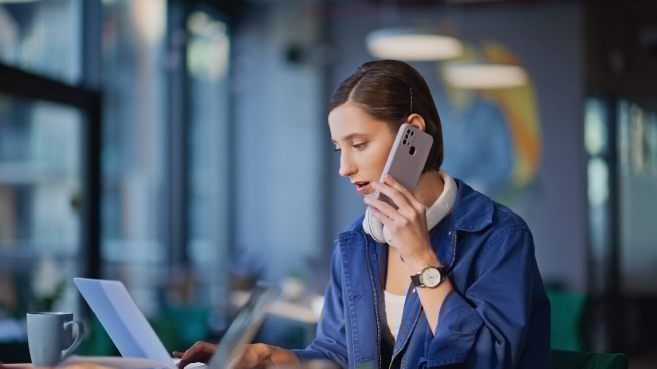 Nuori naispuolinen työntekijä modernissa coworking-toimistossa puhelimessa samalla kun käyttää kannettavaa tietokonetta, kahvikuppi pöydällä ja sumea työtila taustalla.