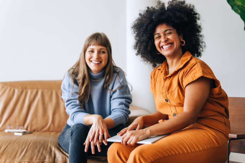 Two successful businesswomen smiling at the camera while sitting together in an office lobby. Two happy female entrepreneurs working as a team in a modern workplace.