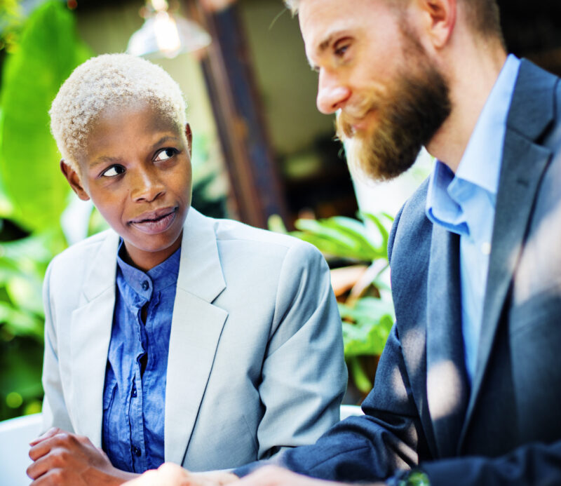 Kaksi ammatillisesti pukeutunutta henkilöä keskustelee istuen; vasemmalla lyhyet vaaleat hiukset ja vaalea bleiseri, oikealla parrakas mies puku päällä, taustalla viherkasveja.