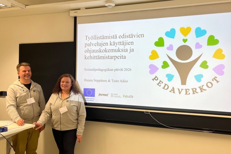 Kaksi henkilöä seisoo luokkahuoneessa esityksen edessä. Taustalla on suuri valkokangas, jossa näkyy otsikko “Työllistämistä edistävien palvelujen käyttäjien ohjauskokemuksia ja kehittämistarpeita” sekä logot ja tapahtuman tiedot.