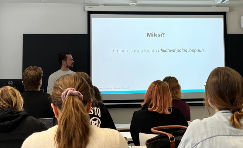 Luokkatilassa pidettävä esitys, jossa yleisö istuu riveissä ja seuraa valkokankaalle heijastettua diaa, jonka otsikko on “Miksi?”. Edessä seisoo yksi henkilö esittelemässä.