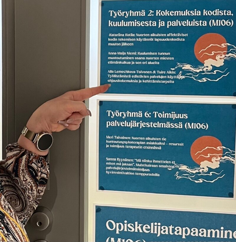 Lähikuva ilmoitustaulusta, jossa näkyy useita työryhmien esittelyjulisteita sinisellä taustalla. Etualalla käsi osoittaa yhtä työryhmäkuvausta.