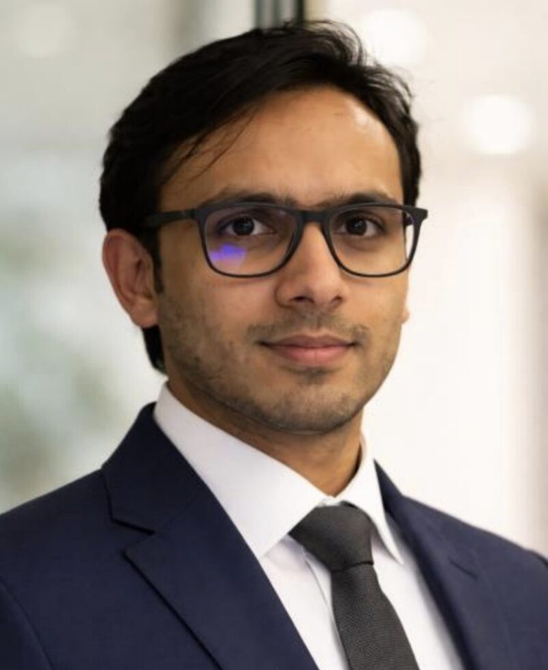 Professional head-and-shoulders portrait of a man wearing dark-framed glasses, a navy suit, white shirt and dark tie, looking directly at the camera with a neutral expression against a soft, out-of-focus light background.