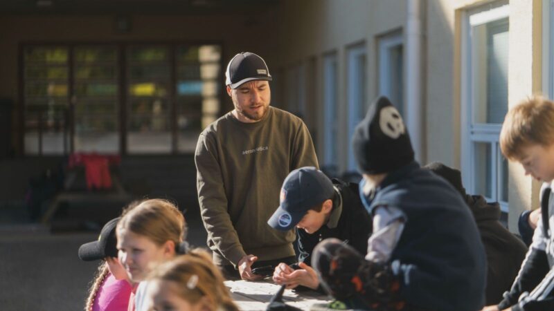Aikuinen mies lippalakissa ohjaamassa useita lapsia pöydän ääressä koulupihalla, lapset kirjoittavat tai piirtävät ja keskustelevat tehtävästä.