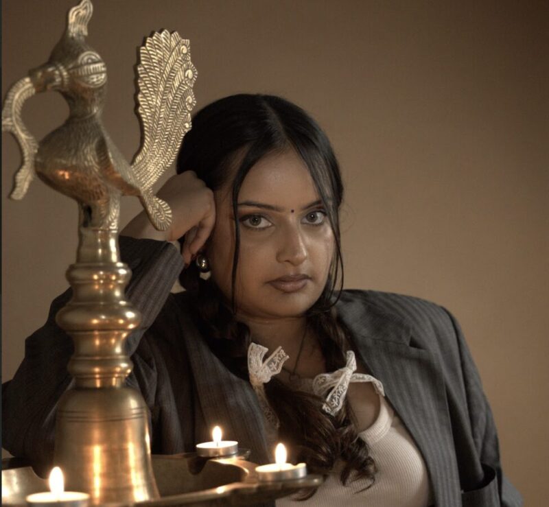 Portrait of a young woman with long dark hair and a neutral expression leaning her arm on an ornate brass oil lamp with several lit diyas, wearing a pinstripe blazer over a light top against a warm brown background with soft, warm lighting.