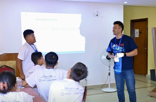 A photo of A photo of Shamsher Tamang smiling in a classroom with five young students. He wears a shirt printed with the text “Use Less Plastic.” A screen with the text “Attendance Sheet” and a rotating fan are in the background. smiling in a classroom with five young students. He wears a shirt printed with the text “Use Less Plastic.” A screen with the text “Attendance Sheet” and a rotating fan are in the background.