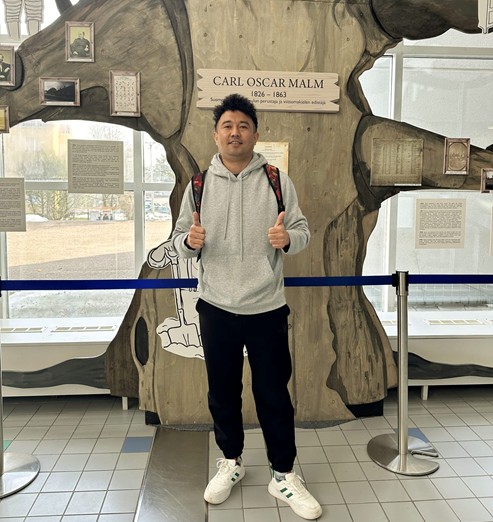 A photograph of Shamsher Tamang standing in front of a tree-shaped display honoring Carl Oscar Malm on Humak’s Haaga campus. Shamsher is smiling, wearing athletic clothes and a backpack, and holding two thumbs up.
