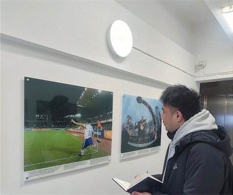 A photograph of Shamsher Tamang, who stands facing to the left. He holds a notebook and looks at images of athletes on a wall at the Olympic Stadium in Helsinki.