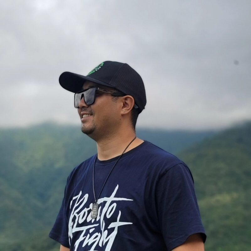 A photo of Shamsher Tamang outdoors, smiling and facing slightly to the left. He wears a T-shirt printed with the text “Born to Fight”, a baseball cap, sunglasses, stud earrings, and a necklace.