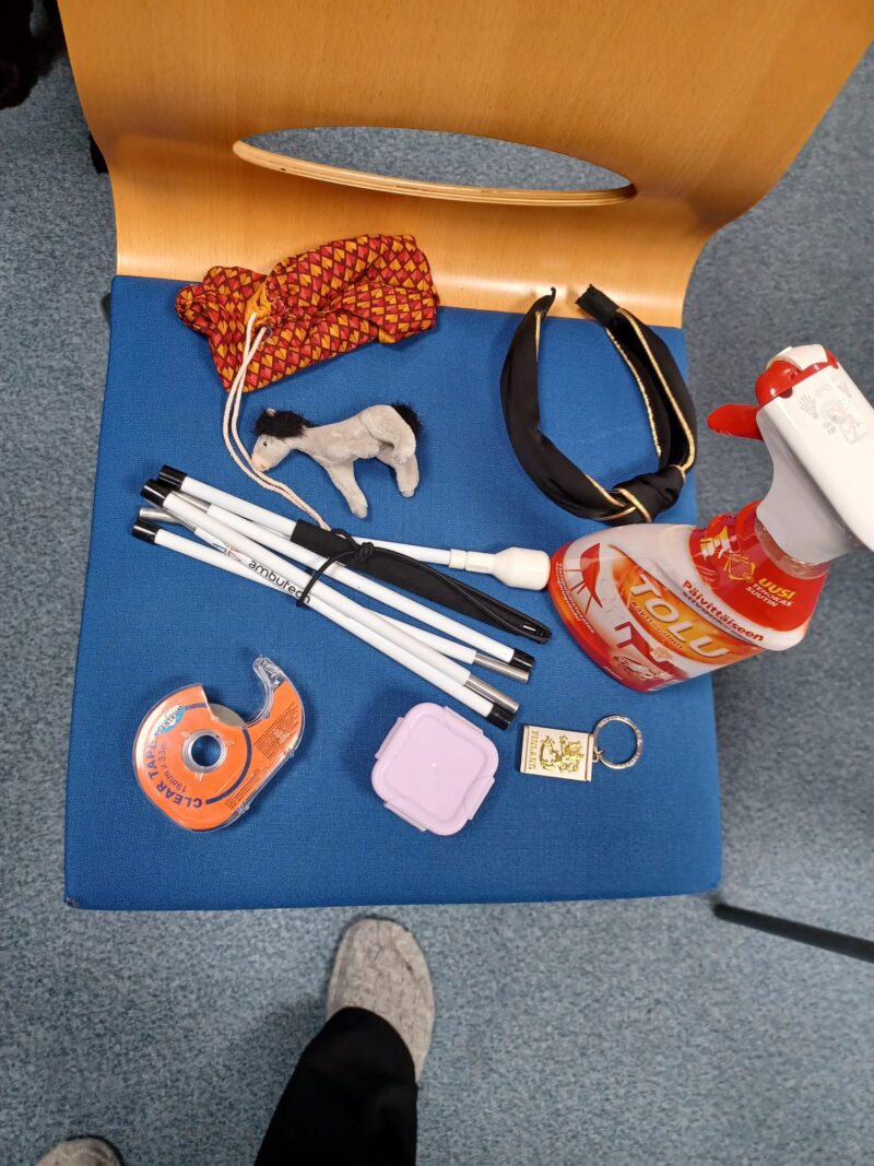 Ylhäältäpäin otettu kuva. Sinisen tuolin päällä on erilaisia esineitä. Oranssi pussukka, musta hiuspanta, valkoinen keppi, pieni harmaa pehmohevonen, punavalkoinen tolusuihkepullo, pieni violetti neliskanttinen muovirasia, hopeinen suomileijona-avaimenperä ja teippirulla.