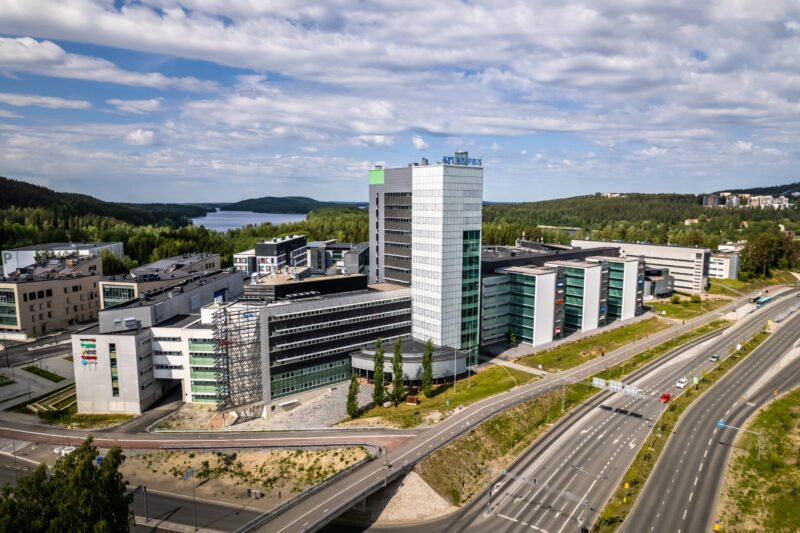 Ilmakuva modernista yrityskampuksesta Kuopiossa: useita toimisto- ja kampusrakennuksia, korkea toimistotorni keskellä, vieressä vilkas tieverkosto sekä metsää ja järvimaisemaa taustalla.