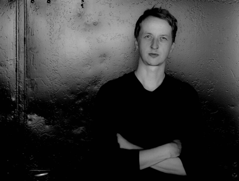 Black-and-white portrait of a young man with short hair wearing a dark shirt, standing with his arms crossed against a textured dark wall and looking directly at the camera with a neutral expression.