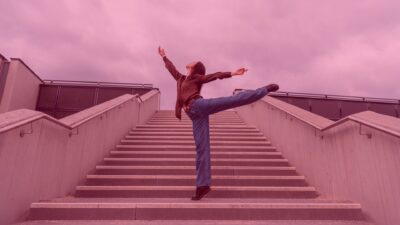 A person stands on wide outdoor stairs, performing a dance-like movement with one leg extended to the side and arms open upward. The sky is pinkish, and the overall image has a soft pink tone.