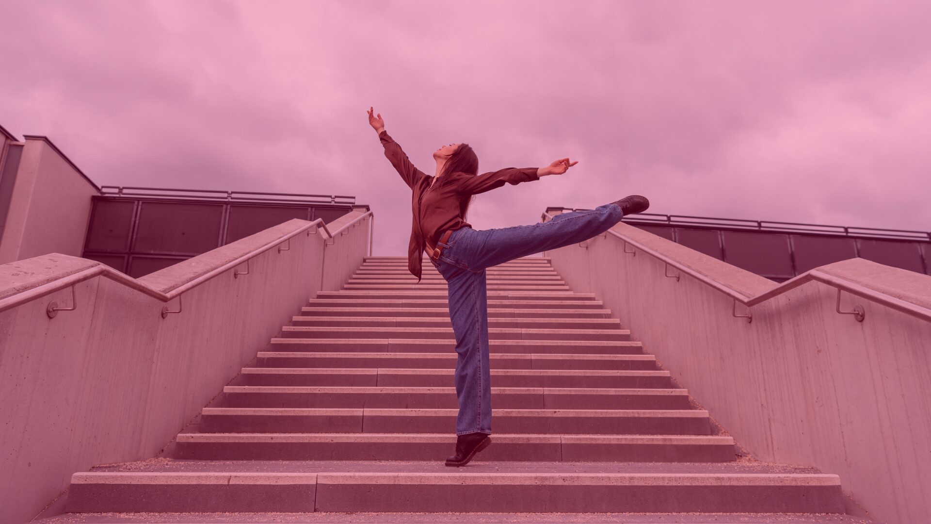 A person stands on wide outdoor stairs, performing a dance-like movement with one leg extended to the side and arms open upward. The sky is pinkish, and the overall image has a soft pink tone.