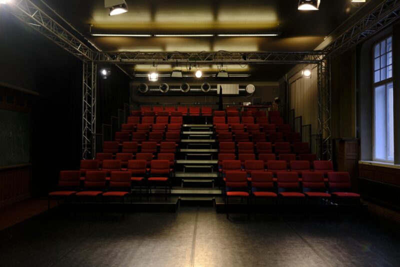 Teatterin lavalta otettu kuva teatterin katsomon penkkiriveistä. Pehmustetut penkit ovat punaisia. Katossa kirkkaat näyttämövalot.