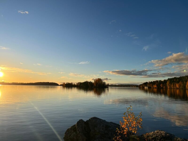 A wide view of a calm Finnish lake at sunset in autumn, with rocky shoreline and one small tree in the foreground. In the background, forested islands and shoreline reflect on smooth water beneath a mostly clear sky with scattered clouds.