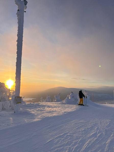 A winter landscape from Lapland at sunset showing a snow-covered hilltop with frost-covered trees and a tall post. A single skier stands near the edge of the slope using ski poles, in front of distant snowy hills.