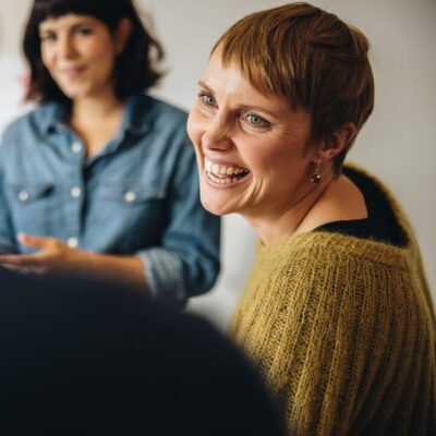 Sisätilassa kaksi henkilöä keskustelussa; etualalla lyhyttukkaisen henkilön hymyilevät kasvot sivuprofiilissa, taustalla toinen henkilö farkkupaidassa, etualalla näkyy kolmannen henkilön hartiaa takaa päin.