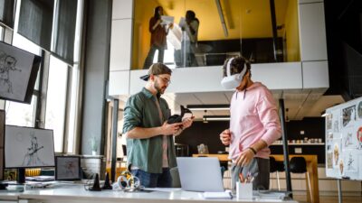 Kaksi henkilöä työskentelee modernissa toimistotilassa: toinen pitää kädessään VR-laseja ja toinen käyttää niitä. Pöydällä on kannettava tietokone ja luonnoksia, ja taustalla näkyy lisää ihmisiä keskustelemassa lasiseinän takana.