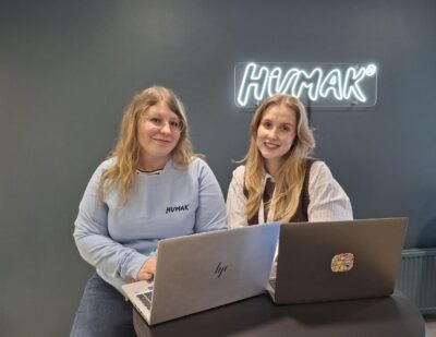 Kaksi henkilöä istuu pöydän ääressä kannettavat tietokoneet edessään sisätilassa; taustalla tummanharmaa seinä ja HUMAK-valo.