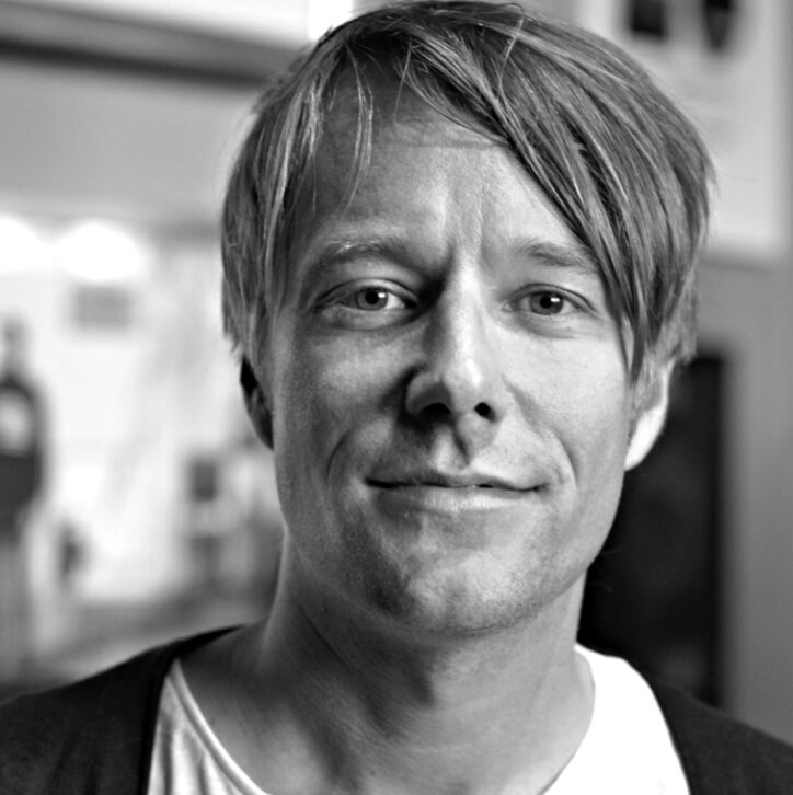 Black-and-white close-up portrait of an adult person with straight hair swept to one side, wearing a light T-shirt and dark cardigan, looking directly at the camera with a slight smile against a softly blurred indoor background.