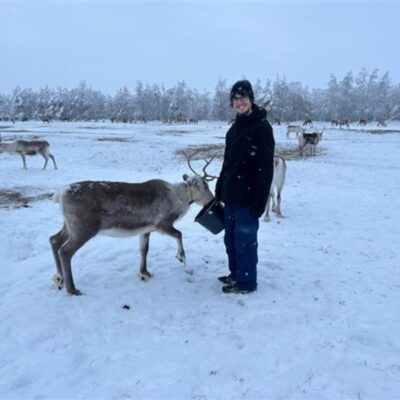 A photo of Hugo in Lapland, smiling and wearing winter clothes. He holds a bucket filled with reindeer food, and one reindeer eats from it. There are snow-covered trees and many reindeer in the background.