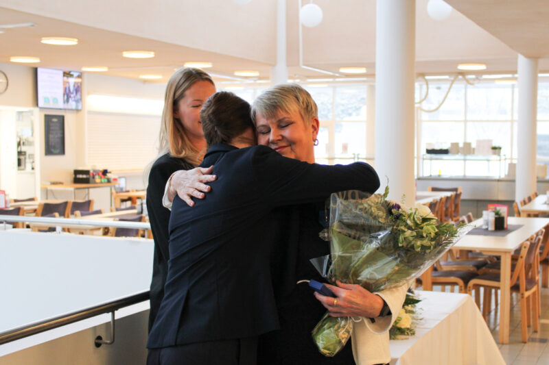 Kuvassa kolme naista, joista kaksi halaa, toinen nainen kuvattu selästä, toinen kasvoista, hymyilee silmät kiinni, kukkaasetelma kädessään, taustalla osin näkyvä nainen ja ravintolasalia.