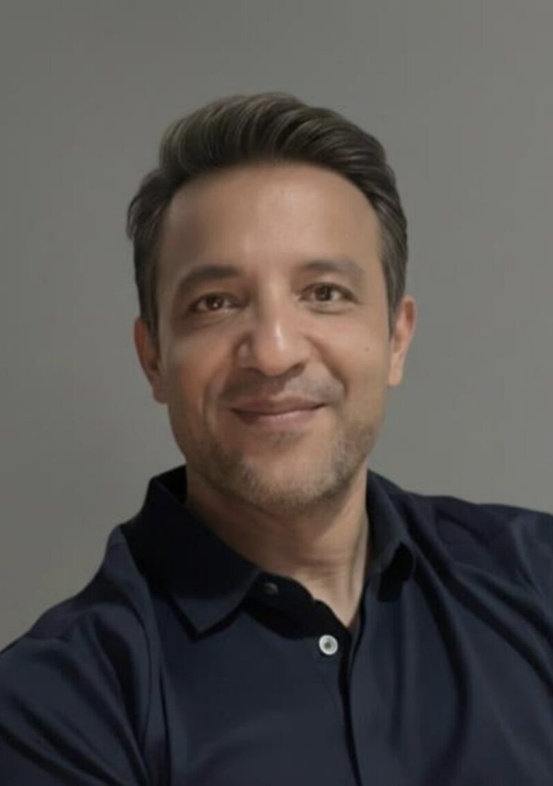 Head-and-shoulders portrait of a smiling middle-aged man with short dark hair and light stubble, wearing a dark collared shirt against a plain gray background.