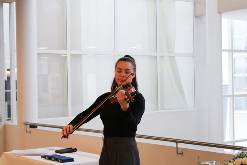Kuvassa nainen soittaa viulua sisätilassa, taustalla isot ikkunat ja parven metallinen kaide.