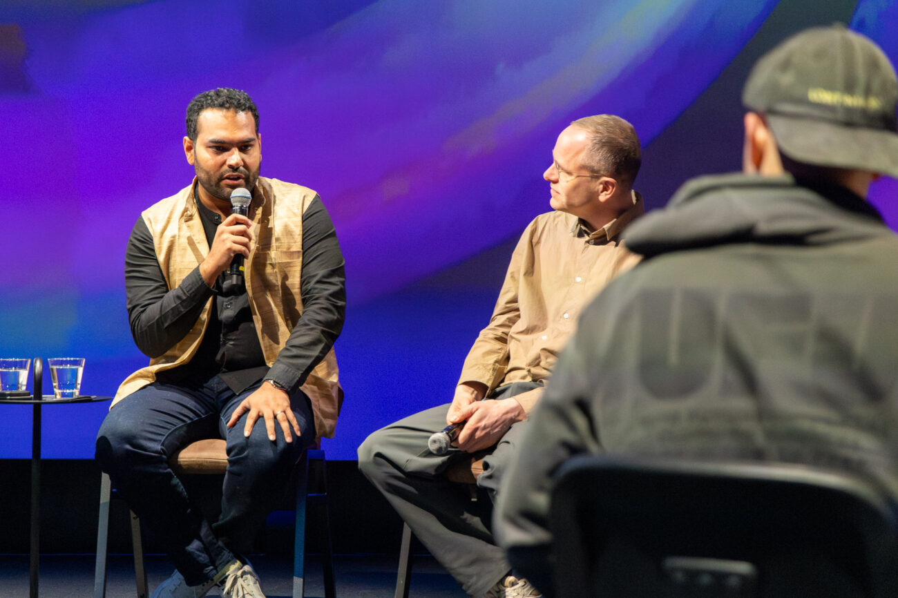 Lavalla kaksi istuvaa henkilöä keskustelutilaisuudessa, molemmilla käsimikrofoni; pienellä pöydällä vesilasit, taustalla violetti–sininen valaistu screen ja etualalla yleisön siluettia.