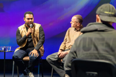 Lavalla kaksi istuvaa henkilöä keskustelutilaisuudessa, molemmilla käsimikrofoni; pienellä pöydällä vesilasit, taustalla violetti–sininen valaistu screen ja etualalla yleisön siluettia.