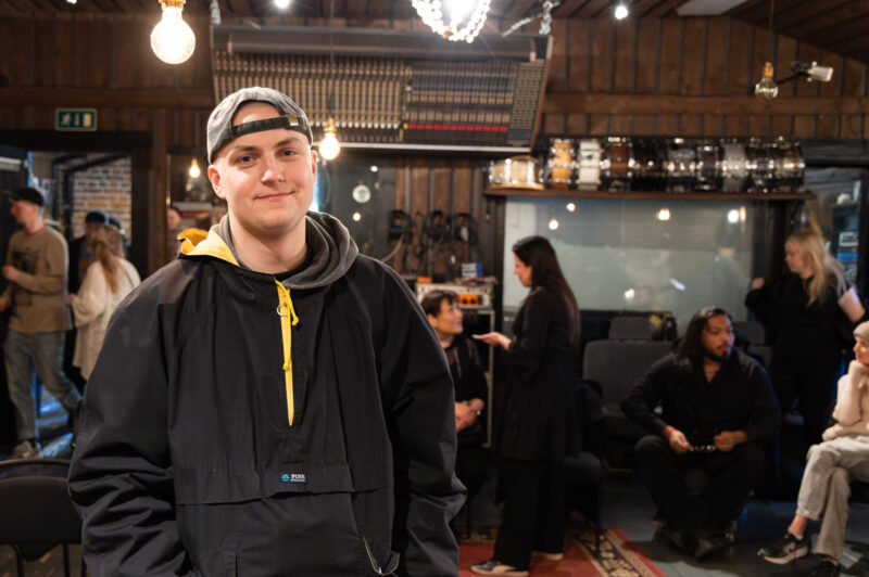 Henkilö seisoo sisätilassa tapahtumapaikalla, yllään tumma takki ja lippalakki. Taustalla näkyy istuvaa yleisöä, tuoleja ja tapahtumatekniikkaa lämpimästi valaistussa tilassa.