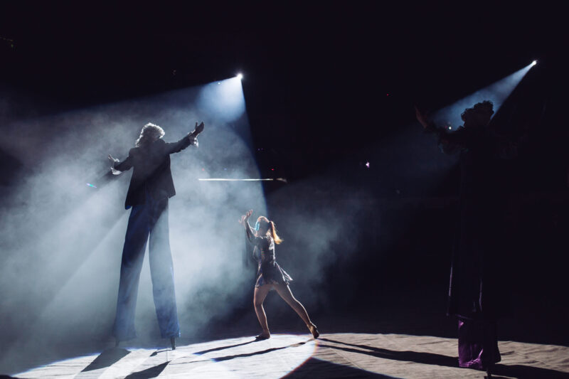 Näyttämökuva esittävästä taiteesta: kolme esiintyjää lavalla voimakkaassa vastavalossa ja teatterisumussa. Keskellä pienempi hahmo liikkeessä, molemmin puolin korkeammalla esiintyjiä kädet kohotettuina. Valokeilat ja varjot korostavat tilaa ja liikettä.
