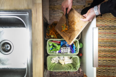 Ylhäältä kuvattu keittiötilanne, jossa henkilön kädet kaapivat leikkuulaudalta kasvisten kuoria ja suikaleita biojäteastiaan tiskialtaan vieressä.