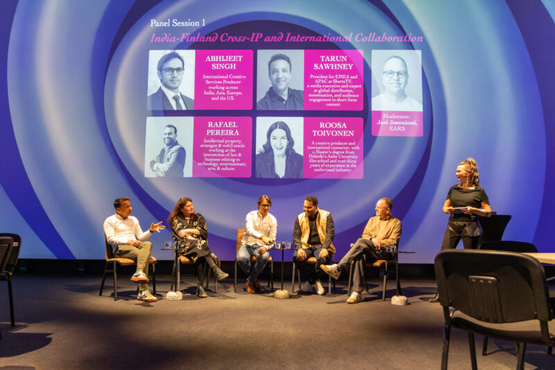 Laaja kuva paneelikeskustelusta: kuusi istuvaa panelistia ja moderaattori lavalla, edessä tuoleja ja pieniä pöytiä vesilaseineen. Taustalla suuri esitysnäyttö, jossa lukee ”Panel Session 1: India–Finland Cross-IP and International Collaboration” sekä osallistujien nimet ja kuvat.