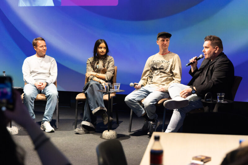 Paneelikeskustelu lavalla: neljä istuvaa panelistia tuoleilla, osalla käsimikrofoni. Edessä pienet sivupöydät vesilaseilla, taustalla sinisävyinen valaistu esitysnäyttö ja etualalla yleisön pöytiä ja tuoleja.