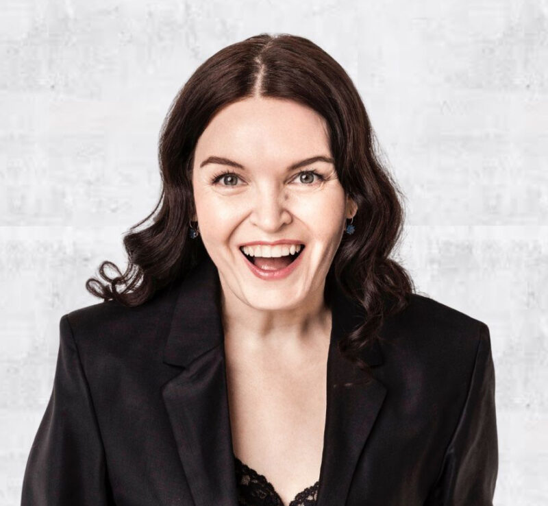 Head-and-shoulders portrait of a woman facing the camera with shoulder-length dark wavy hair and an open, joyful smile, wearing a black blazer over a lace top against a light textured background.