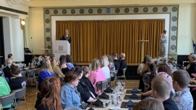 Ryhmäkuva alumnitapaamisesta: kokoussalin täyttyneissä pöydissä istuvat osallistujat katsovat lavaa, jossa vasemmalla puhujapöntöllä henkilö ja oikealla toinen esittäjä lavalla, taustalla suuri keltainen verho ja koristeellinen seinä, pöydillä lasit ja lautasliinat.