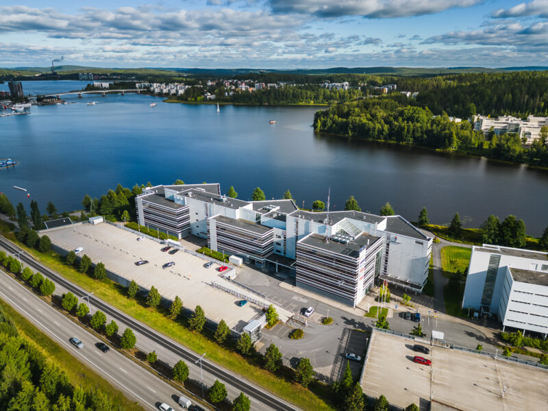 Alt-teksti (suomeksi): Ilmakuva modernista toimistokokonaisuudesta järven rannalla. Etualalla on suuri, monisiipinen toimistorakennus ja laaja parkkialue, vieressä kulkee tie. Taustalla avautuu järvimaisema, silta, veneitä sekä metsäinen rannikko ja kaupunkia rakennuksineen aurinkoisessa säässä.