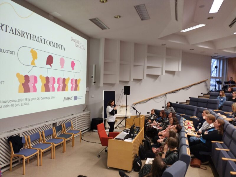 Luentosali, jossa osallistujat istuvat katsomossa kuuntelemassa puhujana seisovaa henkilöä, joka esittää valkokankaalla diaa otsikolla "VERTAISRYHMÄTOIMINTA"; etualalla pöytä, mikrofoni ja rivissä sinisiä tuoleja.