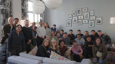 A relaxed, multicultural group of about two dozen young people and adults gathered in a bright living-room, some seated on couches and cushions and others standing behind, smiling for a group photo beneath paper lanterns and a wall of framed photos.