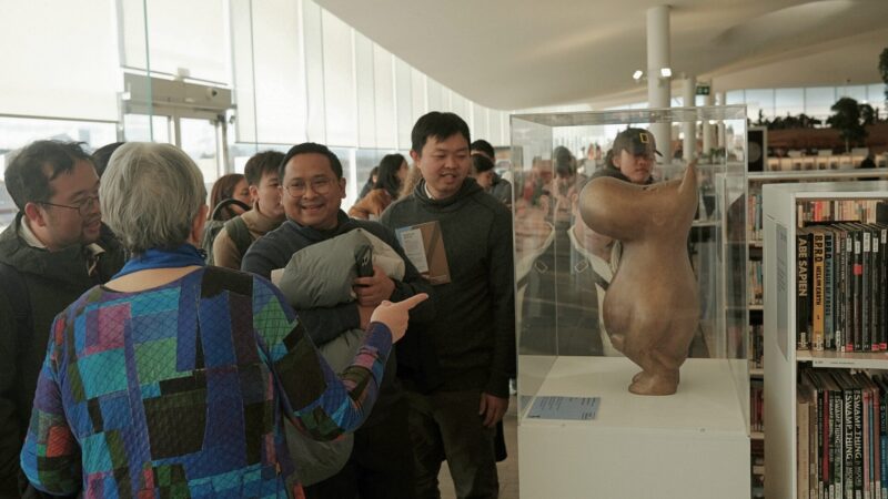 A group of visitors in a bright modern gallery space gather around a glass display case holding an abstract clay sculpture, smiling, pointing and chatting with bookshelves and other exhibits visible in the background.