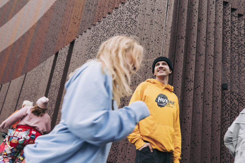 Group of young people outside a tall patterned brick facade; a smiling person in a yellow "Humak" hoodie and black beanie stands center looking up, a blurred blonde person in a light blue hoodie is in the foreground left, and other casually dressed figures are partially visible.