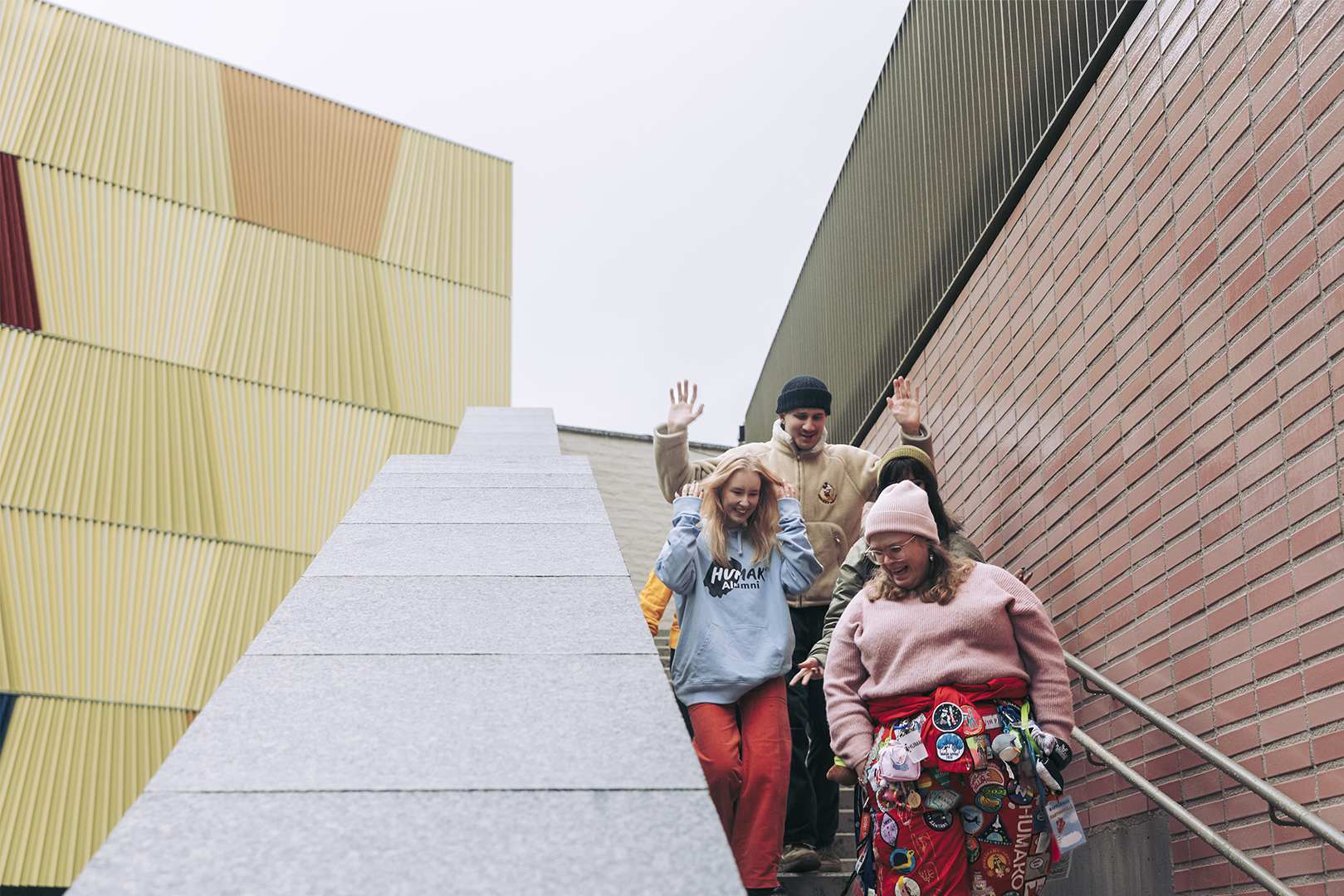 Four smiling people walk down outdoor concrete steps between a brick wall and a yellow modern building, waving and laughing while wearing colorful casual clothing and hats on an overcast day.