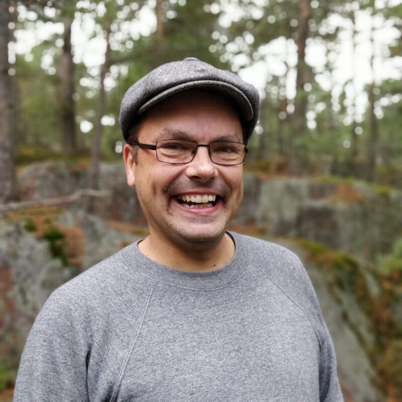 Aikuinen mies hymyilee ulkona metsäisellä kallioisella alueella; hänellä on harmaa lippalakki, silmälasit ja harmaa neule.