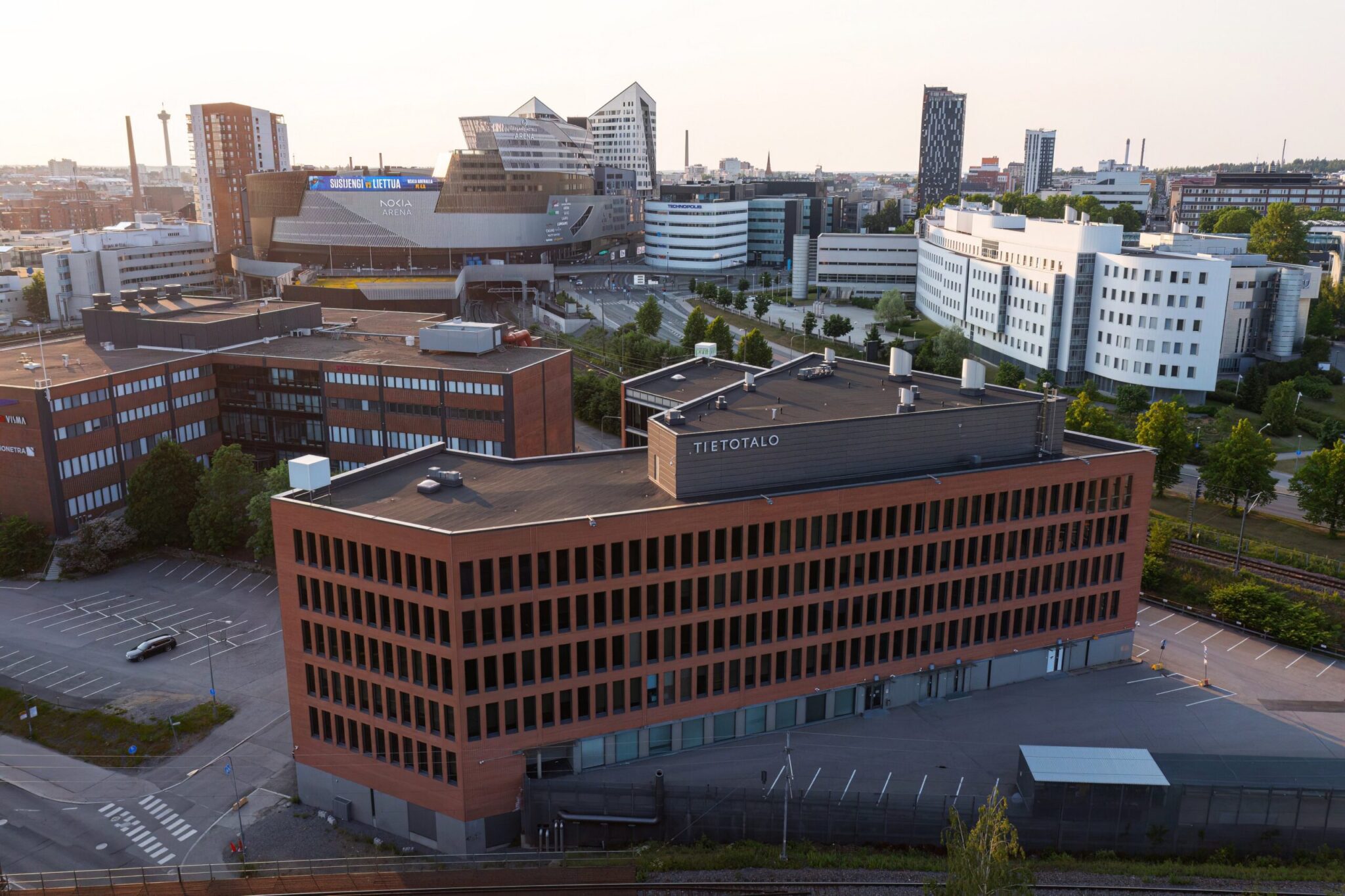 Ilmakuvamainen näkymä, jossa etualalla punatiilinen “TIETOTALO”-toimistorakennus. Taustalla näkyvät Nokia Arena -rakennus, moderneja toimisto- ja tutkimuskiinteistöjä sekä kaupunkimaisema teiden ja rautatien ympäröimänä. Polkuja ja istutuksia kulkee rakennusten välissä aurinkoisessa säässä.