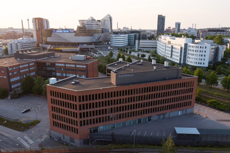 Ilmakuvamainen näkymä, jossa etualalla punatiilinen “TIETOTALO”-toimistorakennus. Taustalla näkyvät Nokia Arena -rakennus, moderneja toimisto- ja tutkimuskiinteistöjä sekä kaupunkimaisema teiden ja rautatien ympäröimänä. Polkuja ja istutuksia kulkee rakennusten välissä aurinkoisessa säässä.