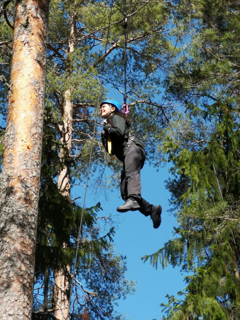 Nuori nainen roikkuu korkealla ilmassa köyden varassa. Ympärillä näkyy puiden latvoja ja taustalla sininen taivas).