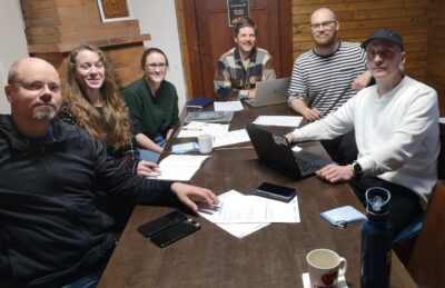 Neljä miestä ja kaksi naista istuu kokouspöydän ympärillä ja katsoo hymyillen kameraan. Kahdella miehellä on kannettava tietokone edessään. Pöydällä on kahvikuppeja ja muistiinpanovälineitä.